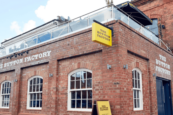 exterior of the button factory with arched factory style windows and red brick. The sign is yellow and there is a glass and metal railing around the top for the roof terrace.
